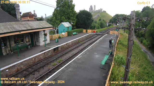 A small train station platform with a stone building on the left side that has a chimney, green benches, and a "Way Out" sign. There are two people near the far end of the platform, engaged in conversation. The railway tracks run parallel to the platform, with a safety fence on the right side. In the background, a lush green hillside features a castle ruin at the top, and there is greenery and trees surrounding the area. An old wooden utility pole with electrical wires runs along the right side of the platform. The scene appears to be early in the morning, as indicated by the timestamp.