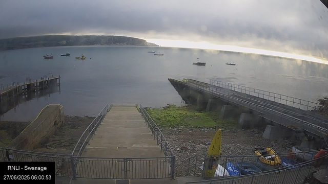 A view of a calm bay with boats anchored in the water. In the foreground, there's a concrete slipway or ramp leading into the water, with metal railings on either side. To the right, a sloped walkway with a railing descends from the top of a small pier or platform. Along the bottom edge, there are yellow and orange safety equipment and some blue storage containers. The sky is overcast with dark clouds, and distant landforms are faintly visible on the horizon. The timestamp shows "27/06/2025 06:00:53" and the label "RNLI - Swanage."
