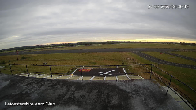 An overcast sky with grey clouds overlooks an airfield with runways and grassy areas. In the foreground, there is a rooftop with a railing, and a model aircraft silhouette painted on the ground below. The timestamp in the upper right corner shows the date as June 27, 2025, and the time as 06:00:49. The bottom left corner has the text "Leicestershire Aero Club."