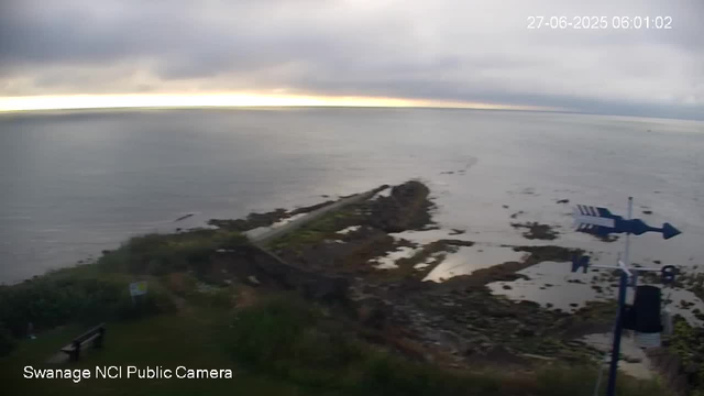 A view of a shoreline with a narrow pier extending into the water, partially covered in grass and rocks. The sky is overcast with gray clouds, and the water appears calm. In the foreground, there is a green area with a bench and a weather vane pointing to the right. The location is identified as "Swanage NCI Public Camera" in the bottom left corner. The timestamp in the upper right corner shows the date as June 27, 2025, and the time as 06:01:02.