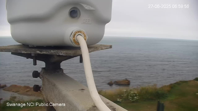 A close-up of a white surveillance camera mounted on a metal pole by the seaside, with the ocean and rocky shoreline visible in the background. The camera is connected with a white cable, and the image includes a timestamp in the top right corner. The bottom left corner displays the label "Swanage NCI Public Camera."