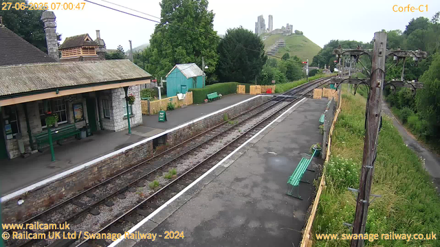 A quiet train station platform in the early morning with two green benches, a small blue shed, and a row of yellow barriers at the far end. There are wooden power poles with electrical wires. In the background, a lush green hill with ruins of an old castle or fortress on top. The sky appears overcast. The timestamp in the top left corner shows 07:00:47 on June 27, 2025.