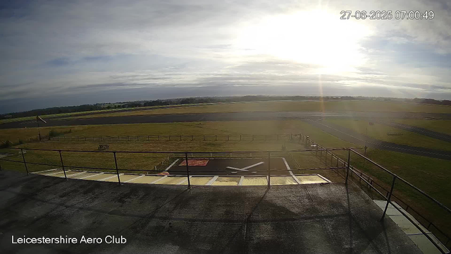 A view from a webcam at Leicester Airport Club showing an open outdoor area with a fence, a small section of runway, and a grassy field extending toward the horizon. The sky is partly cloudy, with the sun shining brightly near the top right corner, creating a glare. The date and time displayed on the image are 27-06-2025 at 07:00:49.