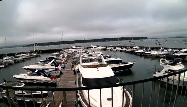 A marina filled with numerous boats and yachts docked along wooden piers under a cloudy sky. The water is calm, and there's a distant landmass with trees on the horizon. The image appears to have been captured from a higher vantage point, possibly from a webcam.