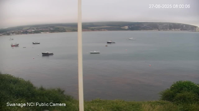 A view of a calm body of water with several small boats floating on the surface. In the foreground, there are green bushes, and in the background, there is a coastal landscape with cliffs and some buildings. The sky is overcast with a layer of gray clouds. The timestamp in the upper right corner shows the date and time as 27-06-2025 08:00:06.