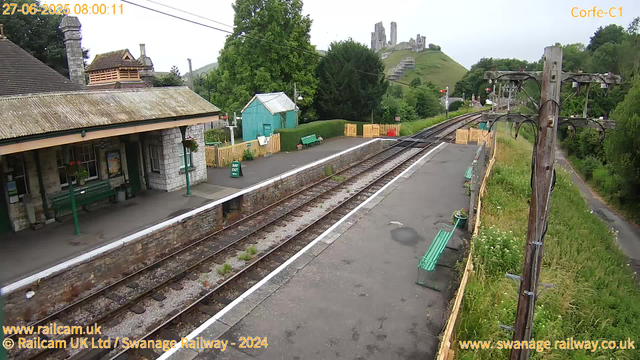 A small, rural railway station platform with a stone building on the left side, featuring a sloped tiled roof, windows, and green benches under the porch. A green sign indicates "Way Out" near a trash bin and a small turquoise shed in the background. There are yellow barriers at the end of the platform, with lush green trees and a hill with ruins or castle remains in the distance. Railway tracks run through the middle, curving toward the right. On the right side, a wooden utility pole with wires extends along the grassy area with wildflowers, and a narrow pathway runs parallel to the tracks. The sky is overcast. The image has a timestamp at the top left corner indicating it was taken on June 27, 2025, at 08:00:11.