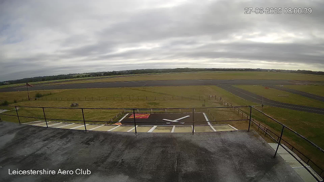 An outdoor view from a webcam at Leicesterhire Aero Club showing a cloudy sky over an airfield. In the foreground, there is a rooftop with a railing and a marked helipad with a large white "H" on a red background. The airfield has runways, grass areas, and some distant structures, with a wide-open landscape extending into the horizon. The timestamp in the top right corner reads "27-06-2025 08:00:39."