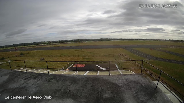 An outdoor view of an airport tarmac and runway under a cloudy sky, with a fenced grassy area in the foreground and a windsock on the left side. The image is timestamped "27-06-2025 08:59:56" and labeled "Leicestershire Aero Club" in the bottom left corner.