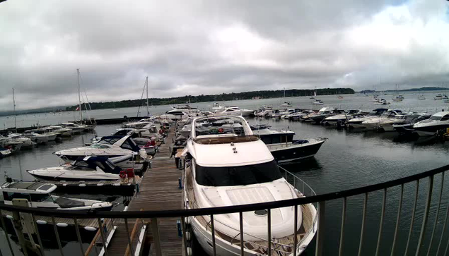 A view of a marina with multiple boats docked along wooden piers. The boats vary in size and style, with some covered and others open. The water is calm, and there is a cloudy sky overhead, with distant land visible across the water.