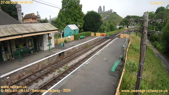 A small station platform with a building on the left side, featuring a sloped roof and stone walls. There are some benches, greenery, and flower baskets on the platform. The railway tracks run through the center, curving slightly to the right as they extend into the distance. A bright green bench is visible on a grassy area beyond the platform, and a small turquoise shed is nearby. In the background, there is a lush green hillside with the ruins of a castle situated atop it. An old wooden pole with electrical wires runs along the right side of the platform, and there is a narrow footpath trail parallel to the tracks on the far right. The scene is bright and appears to be a clear day.