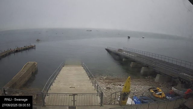 A view of a dock extending into calm water on a foggy day. Several small boats are anchored in the distance. There is a small ramp leading into the water, with a yellow caution sign and some equipment, including yellow and orange life jackets, stored nearby. The scene appears to be at a harbor or seaside location.