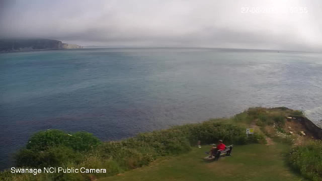 A person wearing a red jacket sits on a bench facing a large body of water, with a grassy area and bushes around them. In the background, there are cliffs along the shoreline under a cloudy sky.