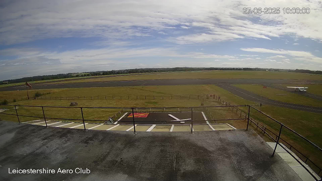 An outdoor view of an airport runway and grassy area taken from a webcam at Leicesterhire Aero Club. The sky is partly cloudy with patches of blue, and the setting appears to be daytime. The foreground shows a concrete balcony or platform with a railing, and a small helipad marked with a white "H" within a red square. In the background, there are taxiways and a hangar or small aircraft to the right. The date and time are displayed in the upper right corner: June 27, 2025, at 10:00 AM.
