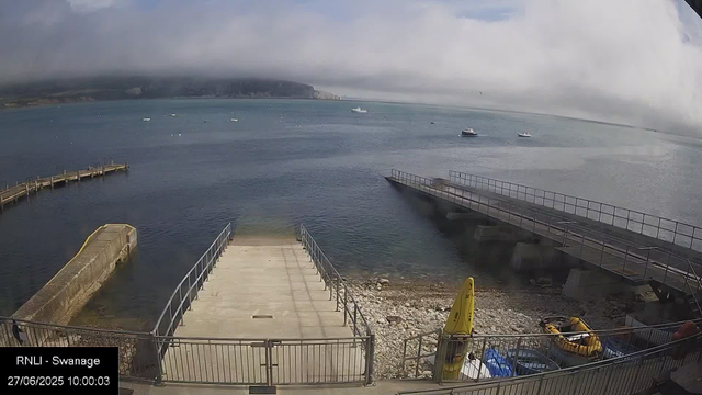 An overcast day at a seaside pier with calm water, several boats in the distance, and a mountain range partially obscured by clouds in the background. The image shows a concrete platform with metal railings, a small yellow boat on a trailer, and some equipment near the dock. The timestamp reads June 27, 2025, at 10:00 am.