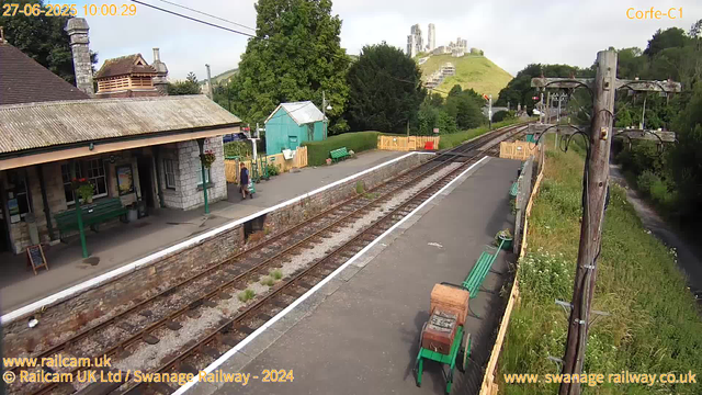 A small rural train station with a single platform, adjacent to railway tracks. There is an old stone building with a tiled roof on the left, featuring green benches and a person standing near the entrance. On the right side, a wooden fence partially encloses the platform, with several green benches and potted plants along the edge. In the background, there are lush green trees, a small turquoise shed, a yellow safety barrier, and a hill with rocky ruins or castle structure at the top. An electrical pole with wires runs alongside the platform. The sky is partly cloudy, and distant hills with a castle or ruins are visible. The image has timestamps and website information overlayed.