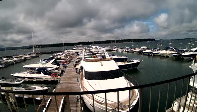 A marina filled with numerous white yachts and boats docked along wooden piers, with a cloudy sky overhead and calm water surrounding the boats.