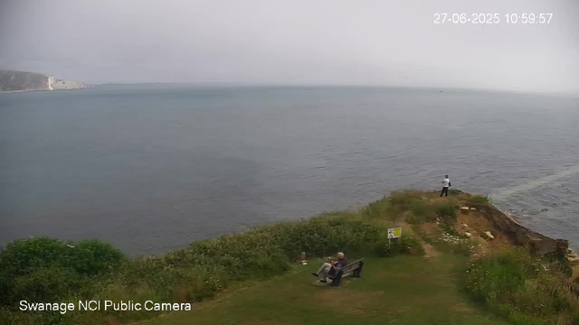 A person is sitting on a bench near the edge of a grassy area, looking out at the water. Another person is standing on the rocky outcrop further ahead, also facing the water. The water is calm, and in the background, there are white cliffs on the distant shoreline under a cloudy sky. The image has a timestamp in the upper right corner and a label at the bottom left that reads "Swanage NCI Public Camera."