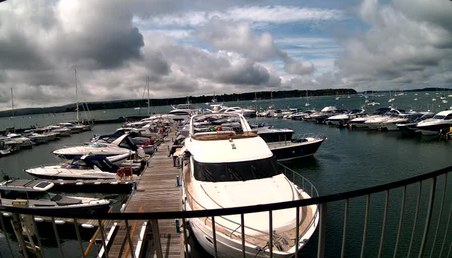 A marina filled with numerous sailboats and motor yachts docked along a wooden pier, with cloudy skies overhead and a distant shoreline visible across the water.