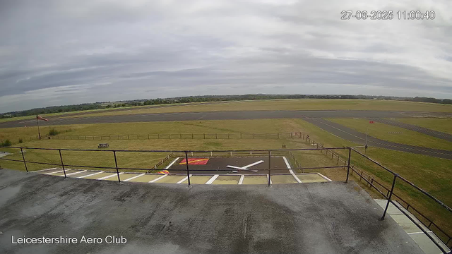 An empty airfield runway view from a webcam at Leicesterhire Aero Club. The sky is overcast with gray clouds, and the runway stretches into the distance with some grassy areas, fences, and a windsock visible in the foreground. The image includes a timestamp of June 27, 2025, at 11:00:40.