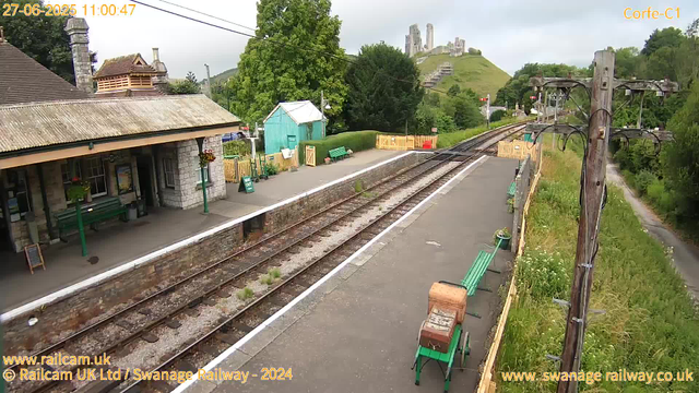 A small train station platform with a stone building on the left, green benches, and flower pots. The train tracks run through the middle, disappearing into the distance. On the right, there are wooden utility poles with wires, and a grassy area with a narrow path leading into trees. In the background, a hillside with old ruins or stone structures atop it is visible under a cloudy sky. The image is timestamped June 27, 2025, at 11:00 AM.