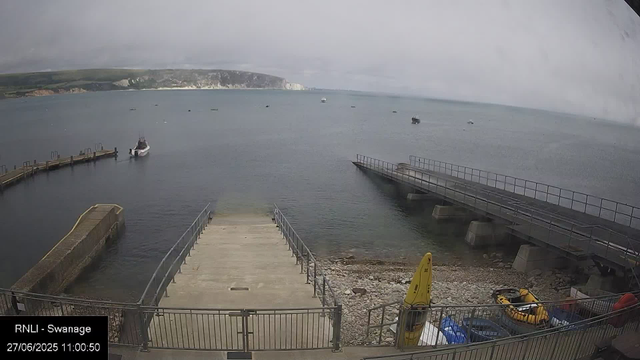 A view of a small harbor with a pier extending into the calm sea. Several boats are anchored in the water, and a rugged coastline with cliffs is visible in the background under a cloudy sky. There is a yellow safety buoy and some equipment near the shoreline in the foreground.