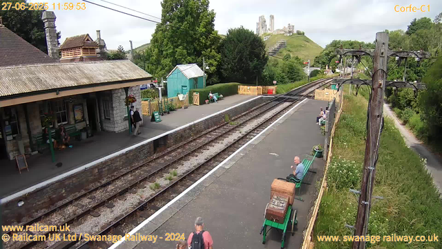 A small train station with a stone building on the left side, with people sitting on green benches under the roof. Some individuals are walking, and one person with a backpack is arriving at the station. There are train tracks running through the middle, with greenery and trees on both sides. In the background, on a hill, there is a partially ruined medieval castle overlooking the scene. On the right side, along the platform, there are more benches with a person sitting on one. A wooden wheel electric pole is on the right side, and a footpath runs parallel behind the vegetation. The sky is partly cloudy. The timestamp on the top left indicates the date is June 27, 2025, and the time is 11:59:53. The image also contains website credits at the bottom.
