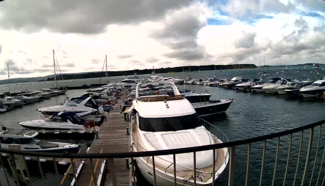 A marina with multiple boats and yachts docked along wooden piers, under a cloudy sky with patches of sunlight breaking through. The foreground shows a black metal railing.