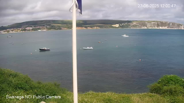 A view of a calm harbor with several boats floating on the water. In the background, there are green hills and white cliffs under a partly cloudy sky. The bottom left corner has the text "Swanage NCI Public Camera." The top right corner displays the date "27-06-2025" and the time "12:00:47."