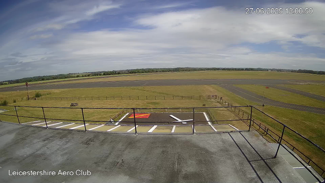 An empty airport taxiway and runway under a partly cloudy sky, with a grassy field surrounding the area. In the foreground, there are parking spaces, a railing, and a small red and white marking on the pavement. The image is timestamped June 27, 2025, at 12:00:50, and labeled "Leicestershire Aero Club" at the bottom left corner.