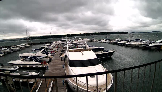 A wooden dock extends into a marina filled with various boats and yachts, all moored closely together. The sky overhead is overcast with dark, heavy clouds, creating a moody atmosphere. In the background, a distant shoreline is visible on the horizon. A black railing runs across the bottom of the image in the foreground.