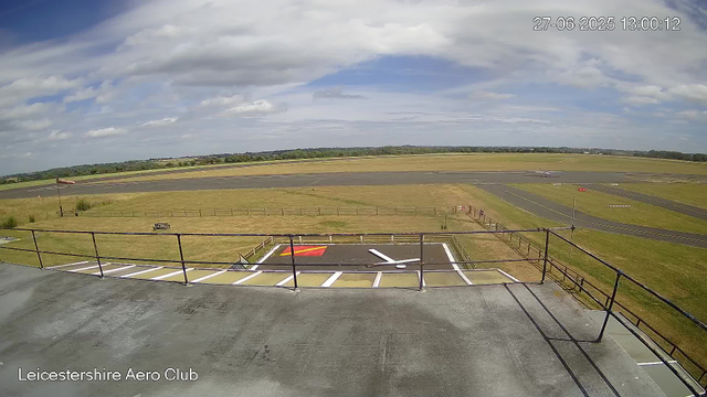 A view from a webcam at Leicesterhire Aero Club showing a runway with some aircraft in the distance, an open grassy area, scattered clouds in the sky, and a small helipad or parking area with a marked helipad in the foreground. The timestamp on the image indicates it was taken on June 27, 2025, at 13:00:12.