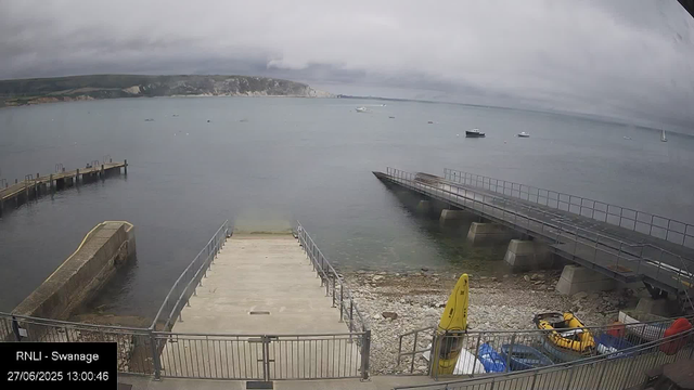 A view of a cloudy day at the waterfront in Swanage, showing a concrete pier extending into calm water, with several boats floating in the distance. There are two jetties or slipways on the shore, one on the left and another on the right, with some yellow safety equipment and a small inflatable boat near the bottom right corner. The background features a hilly coastline under a grey, overcast sky.