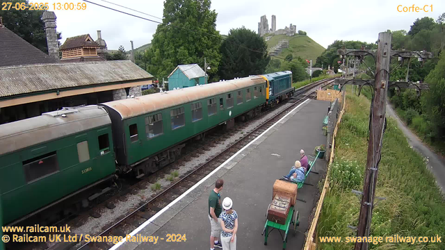 A vintage train with green carriages is stopped at a small railway station platform. Two people, an older man and woman, are seated on a green bench on the platform, engaged in conversation. A young man and a woman, both wearing hats, are standing nearby, talking. An old-fashioned luggage cart is on the platform. In the background, there is a small building with a tiled roof, trees, and a picturesque hill with the ruins of a castle on top. A tall wooden telegraph pole with wires extends along the right side of the platform, and a narrow footpath runs parallel to the railway track. The scene appears to be in a rural area with lush greenery and a scenic landscape. The sky is overcast. The timestamp is "27-06-2025 13:00:59".