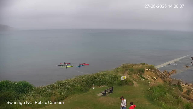 A view of a cloudy day at the seaside with three colorful kayaks (red, yellow, and blue) floating on calm water near the shore. In the foreground, there is green vegetation, a bench, and two people walking along the grassy path, one adult and one child. To the right, there is a rocky coastline with some rocks and small waves hitting the shore. A signboard is also visible among the greenery. The timestamp indicates the date and time as June 27, 2025, at 14:00.