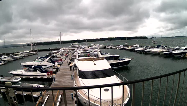 A marina filled with various boats docked along a wooden pier, with a cloudy sky overhead and a body of water extending into the distance.