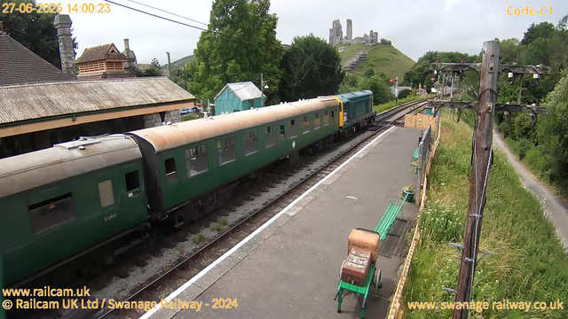 A view of a train station with a green passenger train on the tracks. The train has a weathered, beige roof and dark green body. To the left, there is a building with a tiled roof and chimney. In the background, green trees and a grassy hill with ruins or remains of an old castle can be seen. On the right, there are electrical poles with wires, a narrow path alongside the tracks, green benches, and a small wooden box. The sky is cloudy, and the scene appears to be in a rural or semi-rural area. The timestamp indicates the photo was taken on June 27, 2025, at 14:00.