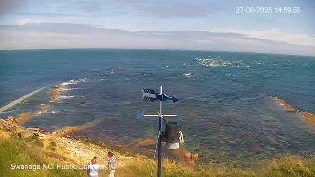A view of a coastal area taken from a webcam, showing the ocean with waves breaking against rocky formations along the shoreline. There are two people standing on a grassy hill near the edge, looking out at the sea. An anemometer and weather instrument are mounted on a pole in the foreground. The sky is partly cloudy with patches of blue, and the timestamp reads "27-06-2025 14:59:53."