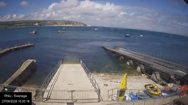 A view of a harbor with several boats floating on the water. In the foreground, there is a concrete pier with railings, a yellow caution sign, and some small boats and vehicles parked nearby. The background features a coastline with cliffs and hills under a partly cloudy sky.
