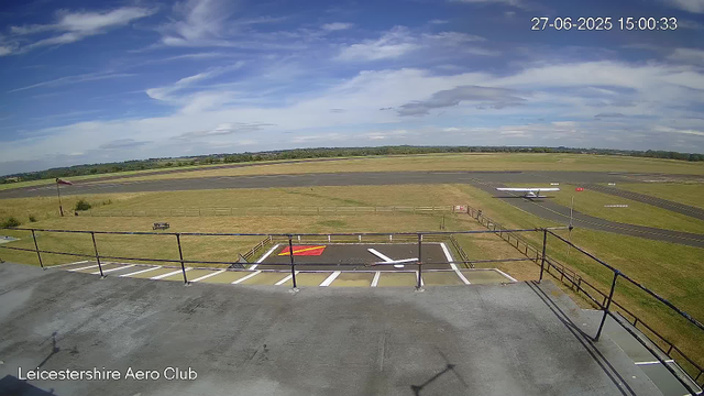 An outdoor view from a webcam at Leicesterhire Aero Club shows a sunny day with scattered clouds in the sky. The scene includes a grassy area in the foreground with a small parked airplane on a runway, and a larger paved runway stretching into the distance. There is a wind sock and a picnic table on the grass, along with fences and markings on the tarmac. The timestamp in the top right corner reads "27-06-2025 15:00:33".