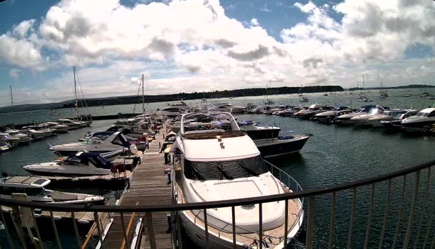 A marina filled with numerous boats and yachts docked along a wooden pier. The sky is partly cloudy with patches of blue visible. The water in the harbor is calm, and the landscape in the distance includes a flat shoreline with some trees.