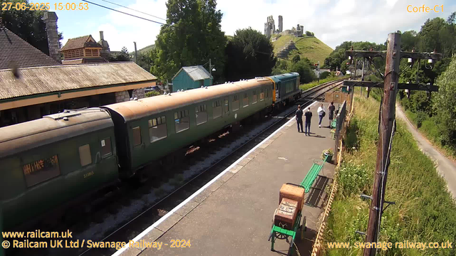 A vintage green train with multiple carriages is stopped at a small rural station platform. Four people are walking along the platform, some carrying bags. To the right, there are wooden utility poles with electrical wires and a small green bench with flower pots nearby. Behind the platform, there are trees, a small building with a sloped roof, and a hill with a castle ruin on top. The sky is partly cloudy with some blue visible. The image is timestamped June 27, 2025, at 15:00:53.