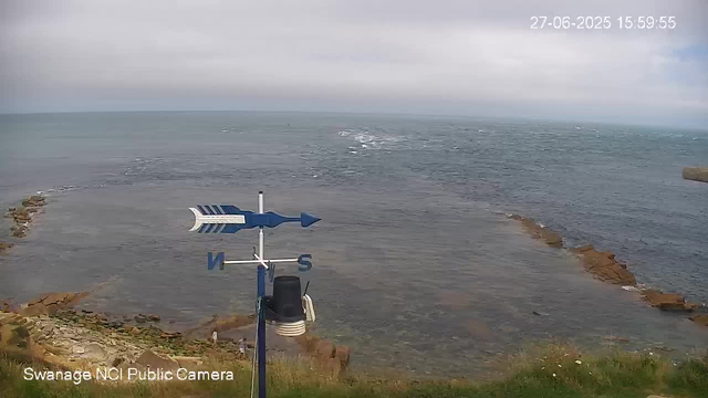 A view of a coastline with a sea and rocky outcroppings, under a cloudy sky. In the foreground, there is a weather vane with a blue arrow pointing east and a black device attached, and a black camera mounted below it. The image has a timestamp in the top right corner showing "27-06-2025 15:59:55" and a label at the bottom left reading "Swanage NCI Public Camera."