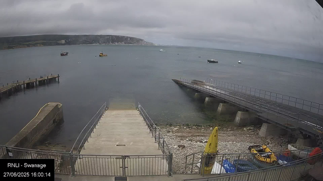 A view of a harbor with three boats floating in the water under cloudy skies. There are two piers extending into the water, one on the left and one on the right, with the right pier having a railing. In the foreground, there is a small dock or platform with a railing, and some equipment including a yellow rescue float, a yellow buoy, and some bags or containers are visible near the bottom right. The background features distant cliffs or hills along the shoreline. The timestamp shows "27/06/2025 16:00:14."