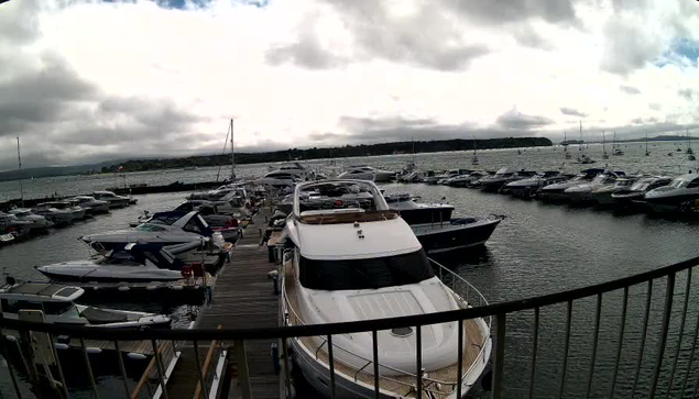 A marina with numerous boats docked along a wooden pier. The boats vary in size and style, with some large yachts and smaller vessels. The water is calm, and there are more boats visible in the distance. The sky above is cloudy with patches of blue peeking through, suggesting an overcast day. A railing runs across the foreground, indicating the photo was taken from a vantage point on a balcony or walkway above the marina.