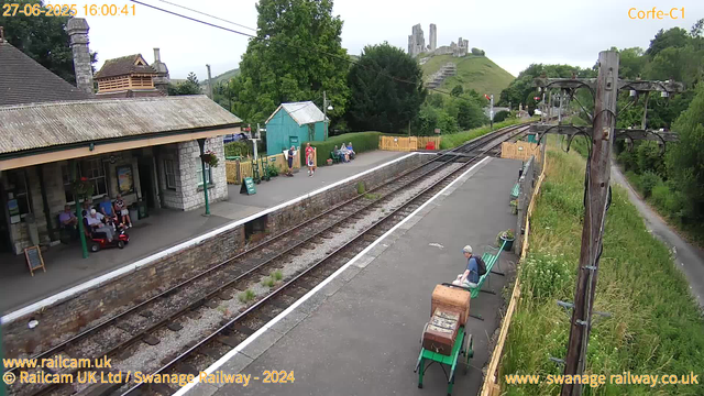 A small railway station platform with a stone building on the left, several people sitting and standing, and a woman walking along the platform. There are green benches, a vintage luggage cart, and a few potted plants. The railway tracks run through the middle of the scene, with a grassy hillside and ruins of a castle on top in the distance. A wooden fence separates the platform from the grassy area, and a utility pole with wires stands on the right side. Trees and greenery surround the station, creating a peaceful rural atmosphere.