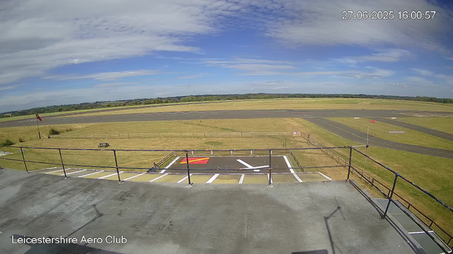 The image shows an outdoor scene at Leicestershire Aero Club, captured from a webcam. In the foreground, there is a parking area with marked spaces, including a designated helicopter parking spot with a red and yellow marking, and a white arrow painted on the ground. A black metal railing runs along the edge of the parking area. Beyond the parking lot, a grassy field extends toward a runway, which is parallel to the horizon. The sky above is mostly blue with scattered clouds, and the scene is well-lit with natural sunlight. The timestamp in the upper right corner indicates the date and time as June 27, 2025, at 4:00 PM.