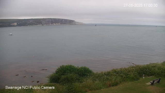 A wide view of a calm sea under a cloudy sky, with a rocky hillside in the background. In the water, there are a few boats and sailboats, some distant. The shoreline has some small rocks, and there is greenery with a bench on the grass in the foreground. The timestamp in the top right corner shows June 27, 2025, at 5:00 PM.