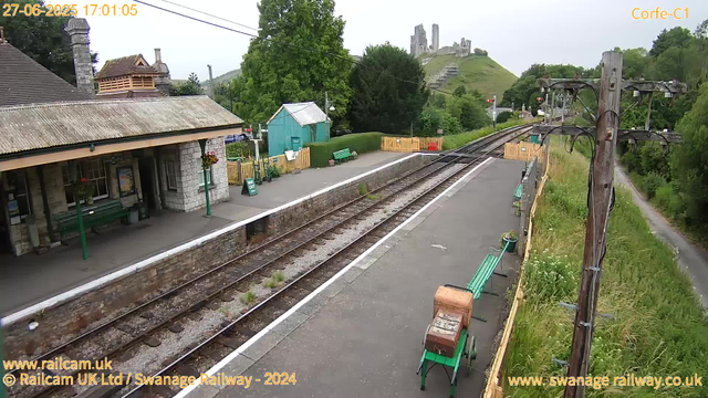 A small railway station platform with a stone building on the left, featuring a sloped roof and a bench outside. There are green benches along the platform, with some flower pots nearby. A yellow fence separates part of the area, and a small turquoise building is in the background. On the right side, there are tall wooden utility poles with wires, and a narrow footpath runs alongside the grass and trees. In the distance, atop a hill, there are ruins of an old castle overlooking the scene. The sky is overcast and the surrounding area is lush with green trees and grass. The timestamp indicates the date and time as 27th June 2025 at 17:01:05.
