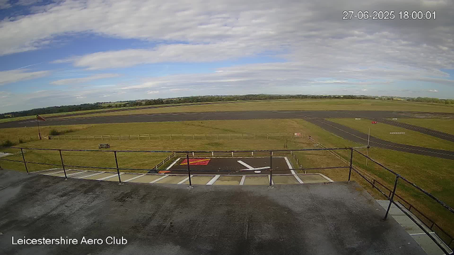 A wide view of an airfield under a partly cloudy sky. In the foreground, there is a small fenced area with a marked helipad, a flagpole with a flag, and some benches. Beyond the fenced area, there are runways and grassy fields extending to the horizon. The timestamp in the top right corner indicates the date and time as June 27, 2025, at 6:00 PM. The bottom left corner displays the text "Leicestershire Aero Club."