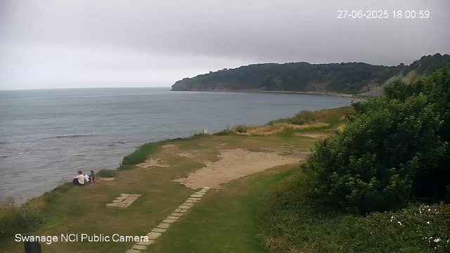 A peaceful coastal scene with a grassy and sandy area leading to the sea. Two people sit near the water's edge on the left side, facing the ocean. There is a large, leafy green bush on the right side, and a distant headland or cliff covered in trees extending into the water in the background. The sky is overcast with dark clouds. The timestamp in the top right corner shows the date and time as June 27, 2025, at 6:00:59 PM. The bottom left corner indicates the image is from "Swanage NCI Public Camera."